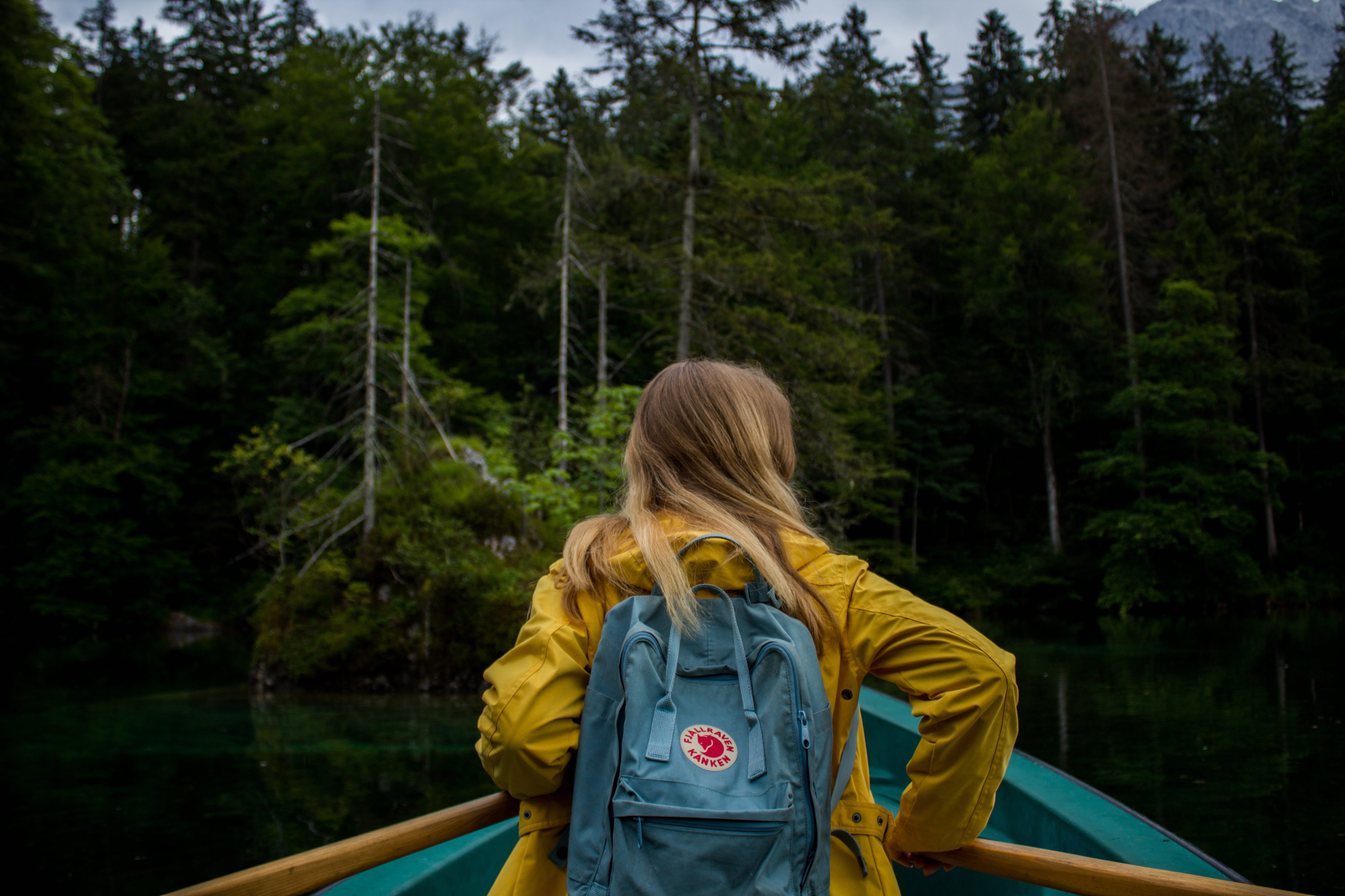 Person in yellow rain jacket with blue backpack sitting by turquoise canoe in lush forest